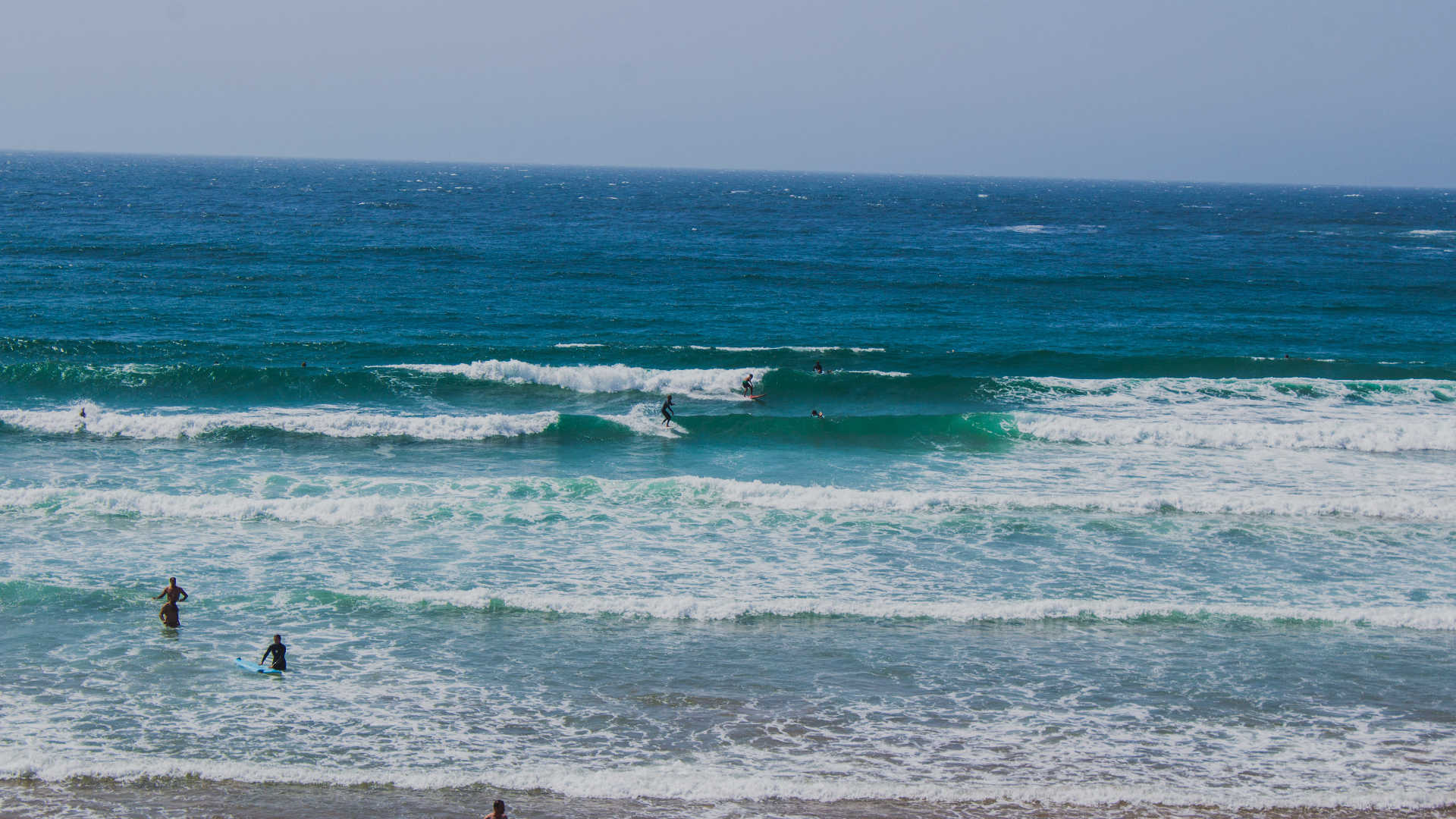 Surfers riding waves in the ocean with people wading in the water at the beach.