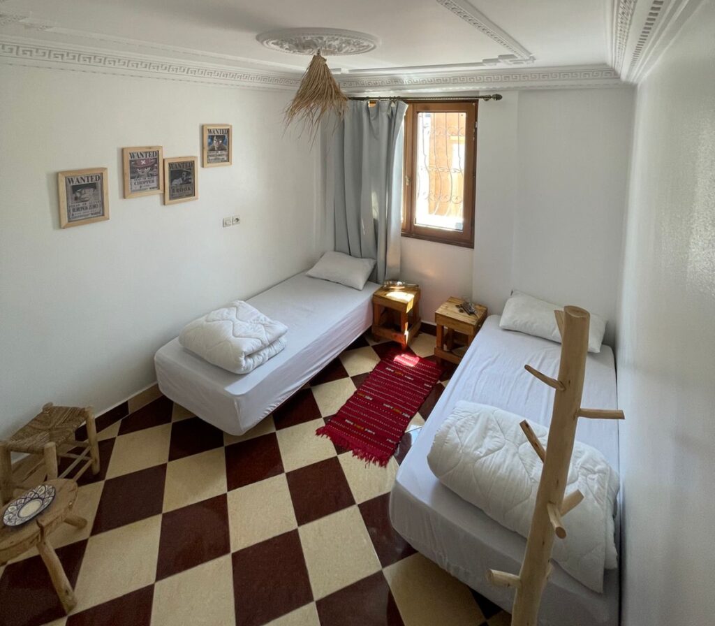 A simple bedroom featuring two single beds, a small table, and wall decorations in a neutral color scheme.